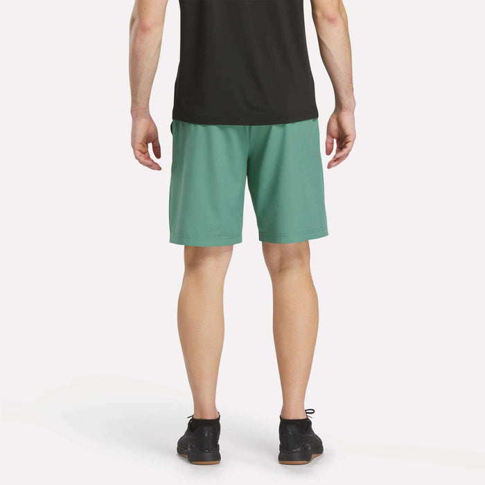 A person stands with their back to the camera, wearing a black shirt, Reebok Men’s Range Woven Shorts 9″ in teal, and black shoes, against a plain white background.