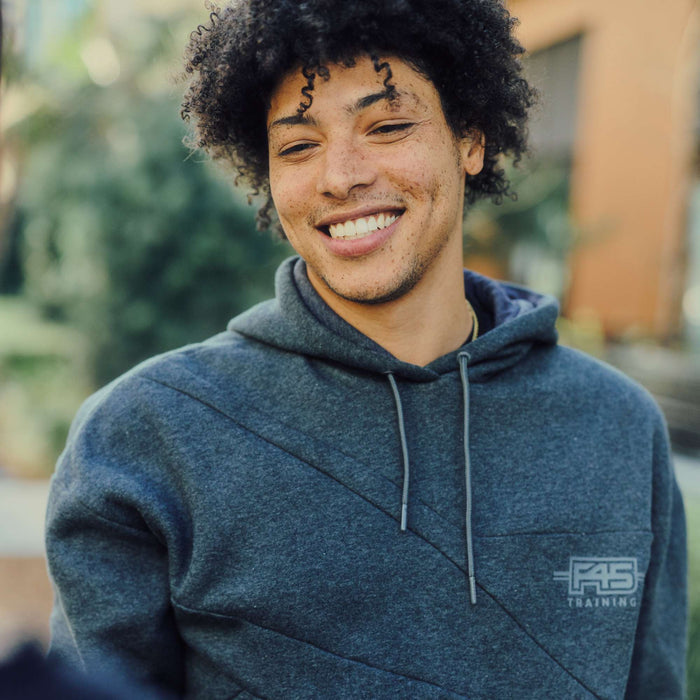 A young man with curly hair and freckles smiles while wearing the Men's Reebok x F45 Training Relaxed Hoodie outdoors, with greenery and a blurred building in the background.