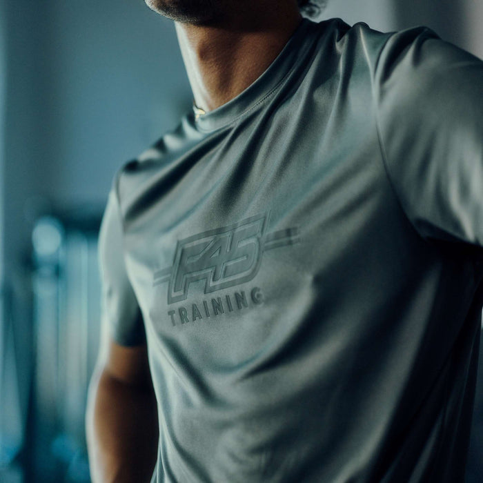 Close-up of a person wearing the Men's Reebok x F45 Training T-Shirt, featuring the Reebok brand logo prominently displayed on the chest. The gray athletic shirt stands out in focus, while a blurred background hints at an indoor gym or fitness environment.