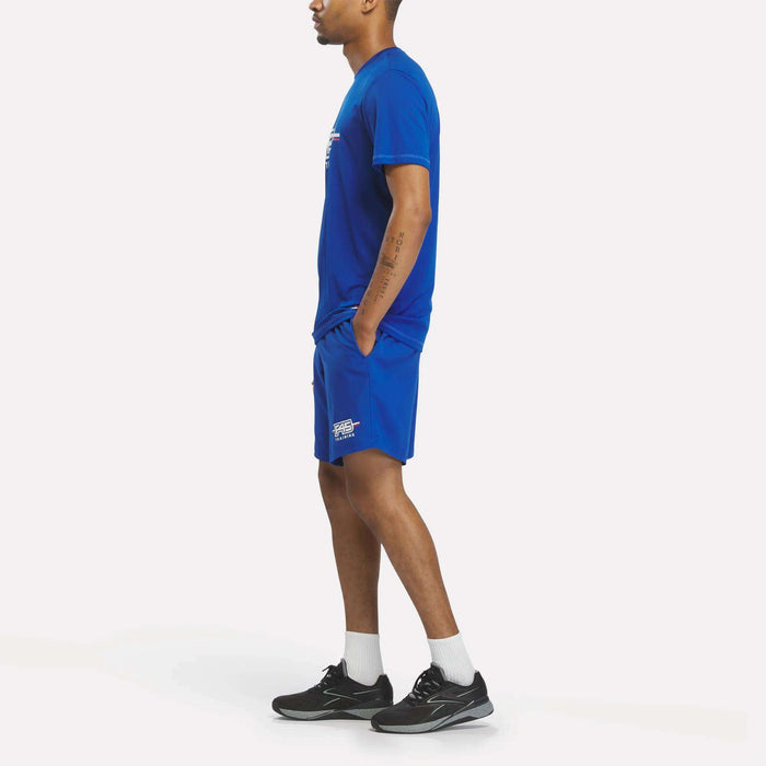 A man stands sideways wearing a Men's Reebok x F45 Training T-Shirt in blue, paired with matching blue shorts, white socks, and black athletic shoes against a plain white background.