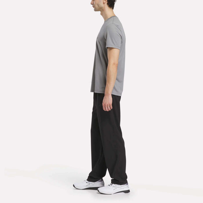 A person stands in profile against a plain white background, wearing black Men's Reebok x F45 Training Woven Pants, a gray T-shirt, and white sneakers—ready for high-intensity workouts or an F45 Training session in this Reebok-inspired look.