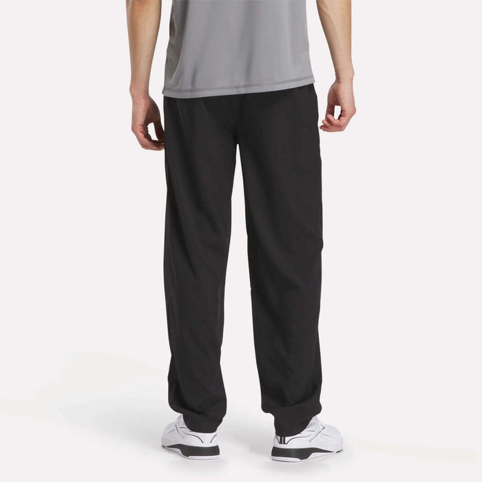 A person stands with their back facing the camera against a plain white background, wearing a gray shirt, white sneakers, and black Men's Reebok x F45 Training Woven Pants from Reebok—an ideal outfit for high-intensity training.