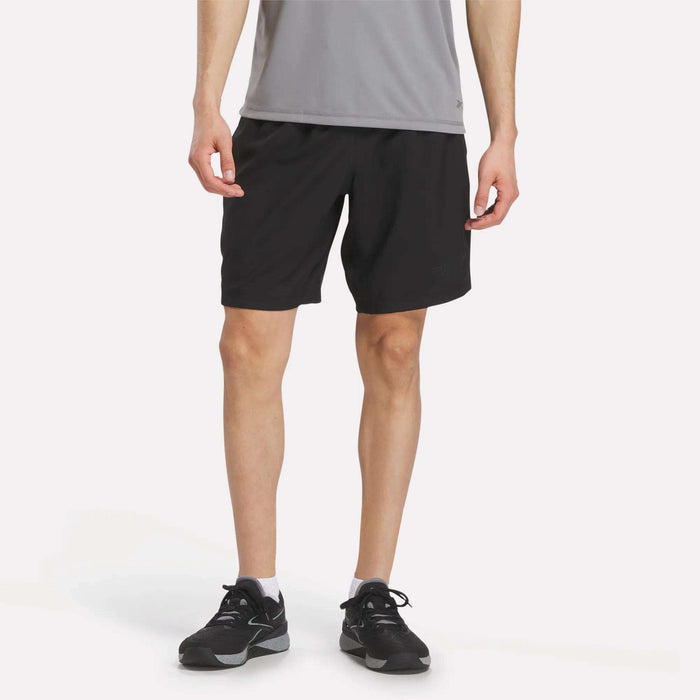 Standing against a plain white background, a person dressed for high-intensity training is wearing a gray shirt, white socks, black sneakers, and black Men's Reebok x F45 Training Woven Shorts. Their head is not visible in the image.