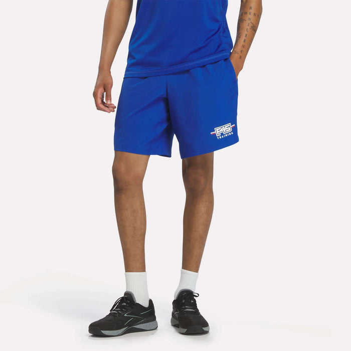 A person wearing a blue t-shirt and Men’s Reebok x F45 Training Woven Shorts in Vector Blue with white logo text, paired with white socks and black athletic shoes, stands against a plain white background—ideal attire for F45 Training or any high-intensity workout.