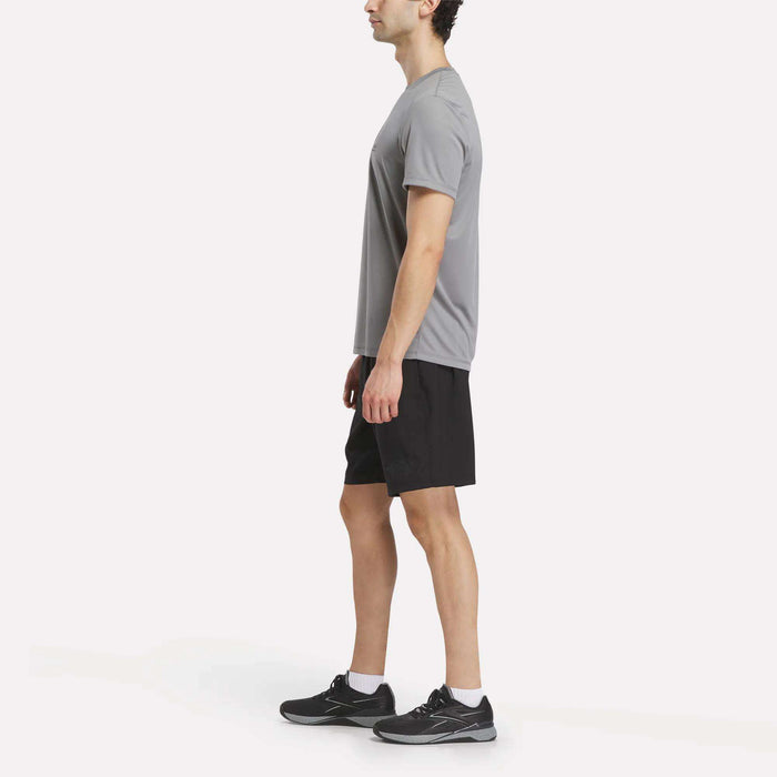 A man stands in profile against a plain white background, dressed for high-intensity training or his next F45 class in a gray T-shirt, black running shoes, ankle socks, and Reebok Men's Reebok x F45 Training Woven Shorts.
