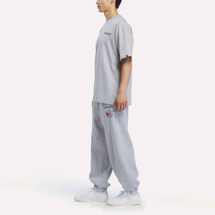 A person in profile wearing the Reebok Men’s Relaxed Logo T-Shirt in gray with matching gray sweatpants featuring small logos, paired with white sneakers, standing against a plain white background.