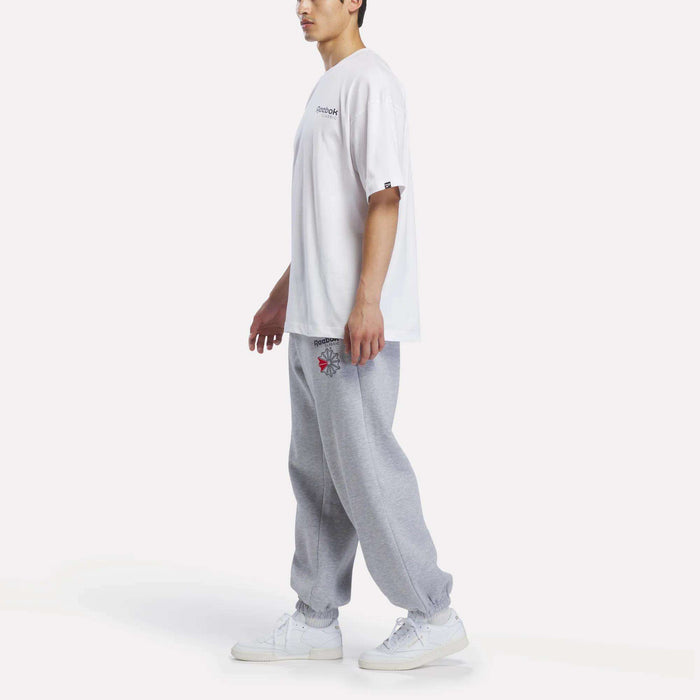 A person wearing a Reebok Men’s Relaxed Logo T-Shirt, gray sweatpants with graphics, and white sneakers stands against a plain white background.