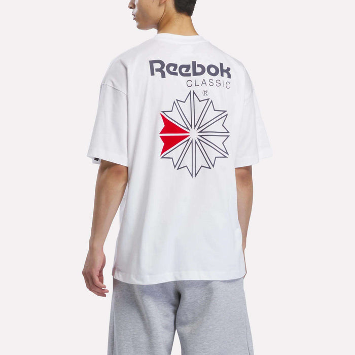 A man is wearing the Reebok Men’s Relaxed Logo T-Shirt—crafted from soft cotton and designed with a bold graphic logo and text on the back—paired with light gray shorts, standing against a plain white background.