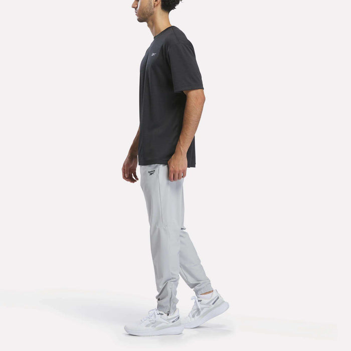 A man stands in profile wearing a black t-shirt, Reebok Men’s Repeat Pants in light gray, and white sneakers against a plain white background.