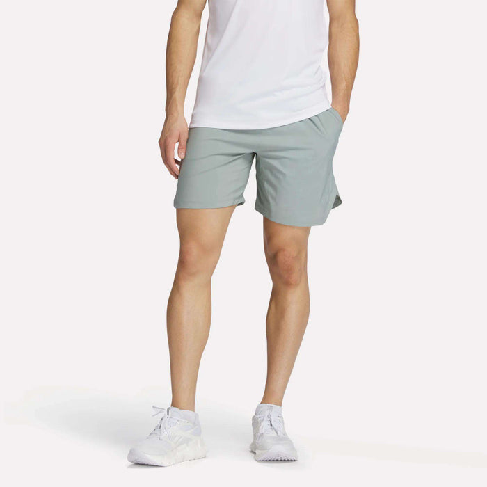 A man stands with one hand in his pocket, wearing a white t-shirt, Reebok Men's Run Shorts with inner tights in light gray, and white sneakers against a plain background.