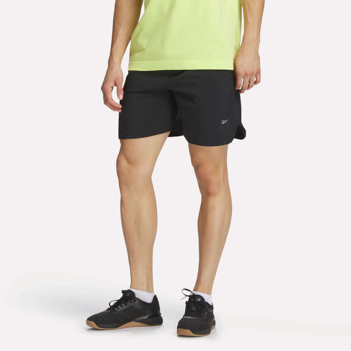 A person in a light green shirt, black Reebok Men's Run Shorts with inner tights, black athletic shoes, and white socks stands against a plain white background. The person's head is not visible in the image.