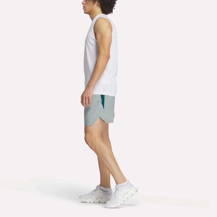 A person in a white sleeveless top, Reebok Men's Run Shorts with inner tights, and white sneakers stands sideways against a plain white background.