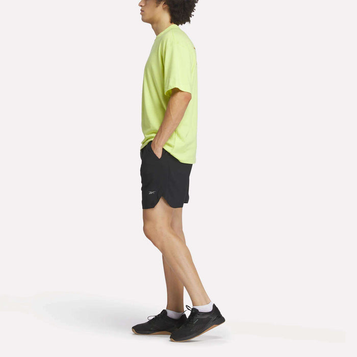 A person stands in profile against a plain white background, wearing a bright yellow T-shirt and Reebok Men's Run Shorts with inner tights, paired with black running shoes and white socks.
