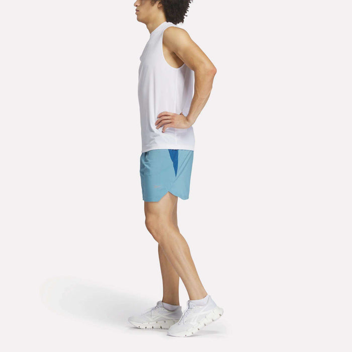 A person stands in profile with hands on hips against a plain white background, wearing Reebok Men's Run Shorts paired with a white sleeveless shirt and white sneakers.