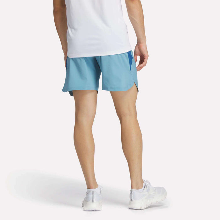 A person stands facing away, wearing a white shirt, Reebok Men's Run Shorts in light blue with inner tights, and white running shoes against a plain light background.