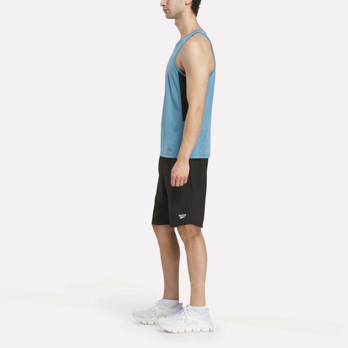 A man stands in profile against a plain white background wearing the Reebok Men's Run Speedwick Singlet in blue, paired with black shorts and white sneakers.