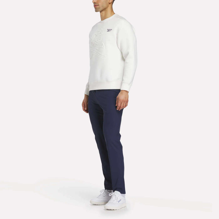 A man stands slightly to the side, wearing a white Reebok Men's Scuba Knit Crew Sweatshirt, navy blue pants, and white sneakers against a plain white background.