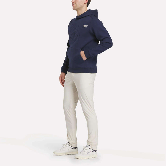 A person stands against a plain background, wearing a navy blue Men's Scuba Knit Hoodie Sweatshirt by Reebok, light beige pants, and white sneakers, with hands in the hoodie pocket and looking slightly to the side.