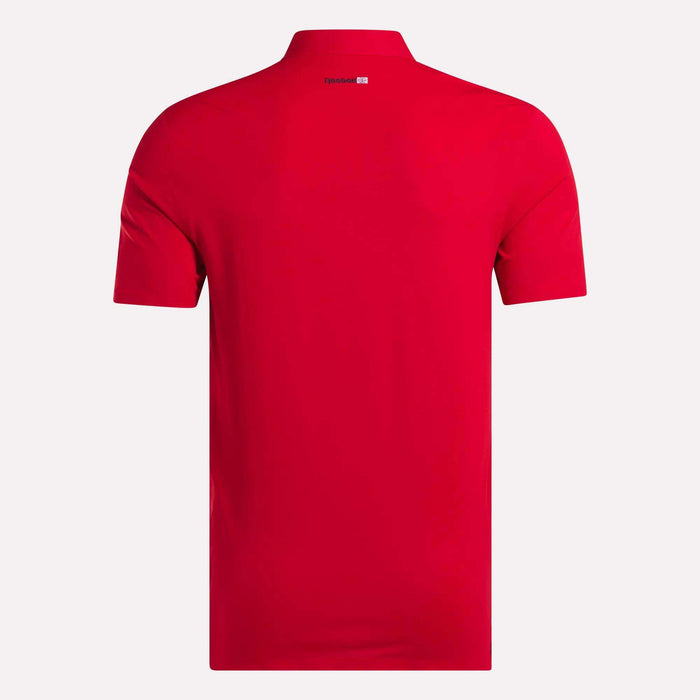 A men's Reebok Short Sleeve Bonded Micro Pique Polo Shirt in plain red is shown from the back, with a small white and red logo by the collar. Made with moisture-wicking fabric, it’s set against a simple light background.