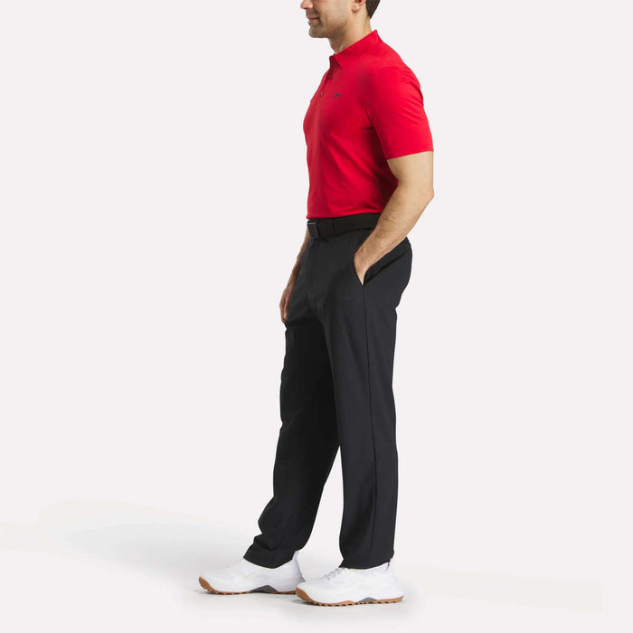 A man stands in side profile with hands in pockets, wearing a red Reebok Men's Short Sleeve Bonded Micro Pique Polo Shirt, black pants, white sneakers, and a black belt against a plain white background.