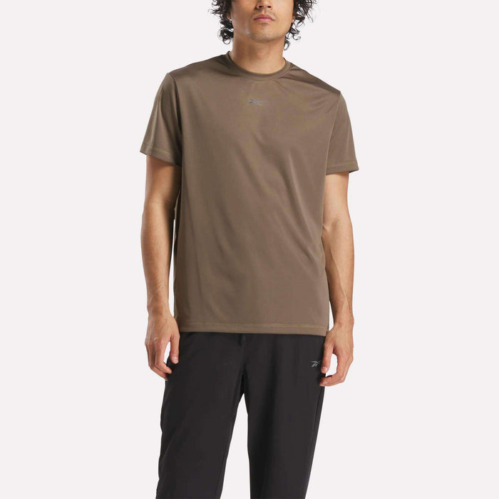 A man with curly hair wears the Reebok Men's ID Train Short Sleeve Tech Tee in brown and black pants, standing against a plain white background.