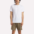 A man stands facing forward wearing a plain white Reebok Men's ID Train Short Sleeve Tech Tee and olive green shorts against a light background. His relaxed stance highlights the sweat-wicking comfort of this classic gym shirt.