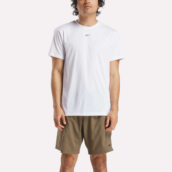 A man stands facing forward wearing a plain white Reebok Men's ID Train Short Sleeve Tech Tee and olive green shorts against a light background. His relaxed stance highlights the sweat-wicking comfort of this classic gym shirt.