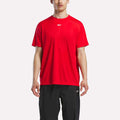 A man wears a Reebok Men's Short Sleeve Tech Training T-Shirt in bright red and black pants, standing against a plain white background. Only his upper body and upper legs are visible.