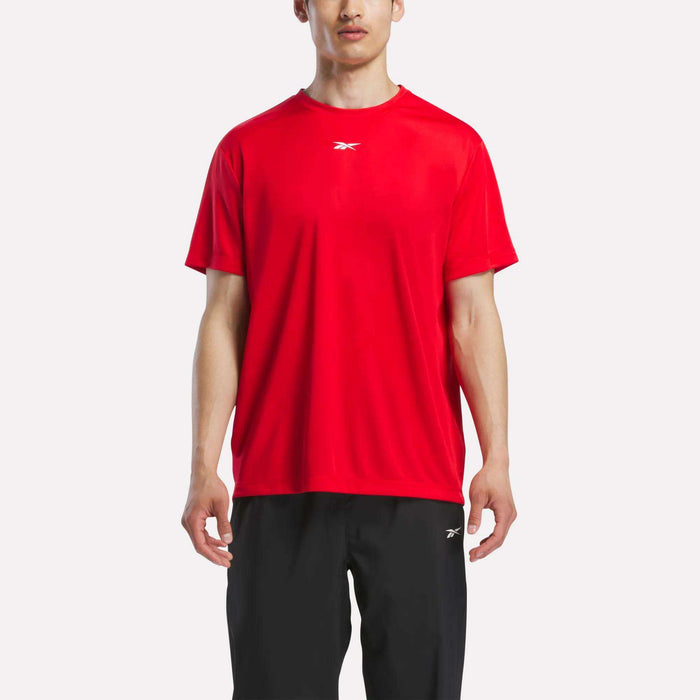A man wears a Reebok Men's Short Sleeve Tech Training T-Shirt in bright red and black pants, standing against a plain white background. Only his upper body and upper legs are visible.
