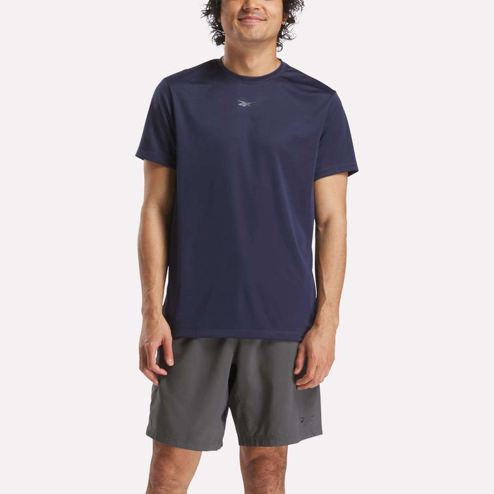 A man stands smiling, wearing a dark blue Reebok Men's ID Train Short Sleeve Tech Tee and gray shorts against a plain light background.