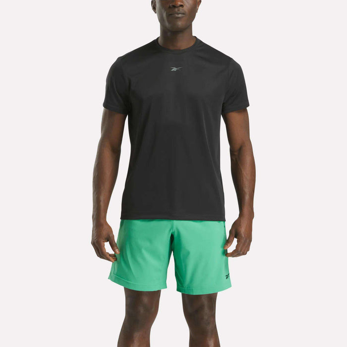 A man wearing a black Reebok Men's ID Train Short Sleeve Tech Tee and light green shorts stands facing forward against a plain white background.