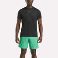 A man wearing a black Reebok Men's ID Train Short Sleeve Tech Tee and light green shorts stands facing forward against a plain white background.