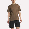 A man stands against a white background wearing the Reebok Men's Short Sleeve Tech Training T-Shirt in brown with a small white chest logo and black shorts.