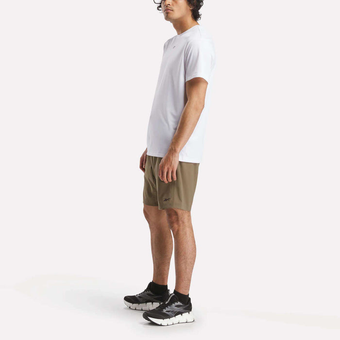A man stands against a white background wearing the Reebok Men's ID Train Short Sleeve Tech Tee, olive green shorts, black socks, and black running shoes. He faces slightly sideways with his arms by his sides.
