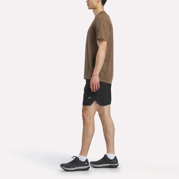 A person stands sideways in a Reebok Men's Short Sleeve Tech Training T-Shirt, black shorts, white socks, and black athletic shoes against a plain white background.