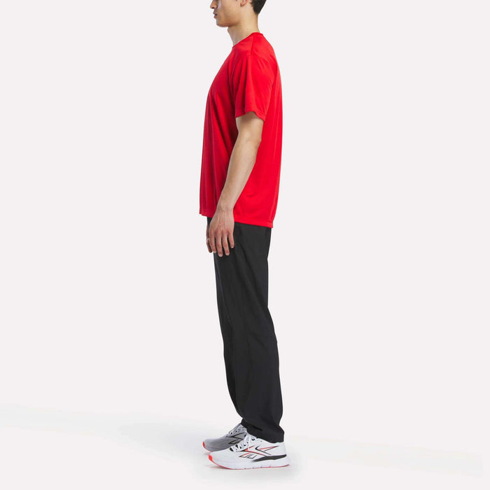 A person stands in profile facing left, wearing a red Reebok Men's Short Sleeve Tech Training T-Shirt, black pants, and white athletic shoes against a plain white background.