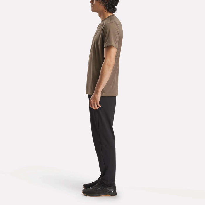 A person stands in profile facing left, wearing a brown Reebok Men's ID Train Short Sleeve Tech Tee, black pants, and black shoes against a plain white background.