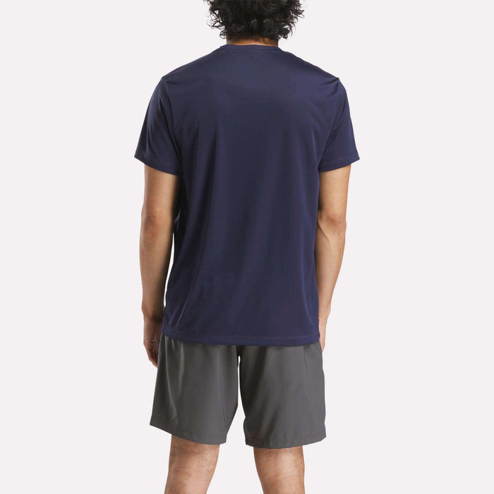 A person with short curly hair, seen from behind, wears a dark navy Reebok Men's ID Train Short Sleeve Tech Tee and gray athletic shorts against a plain white background.