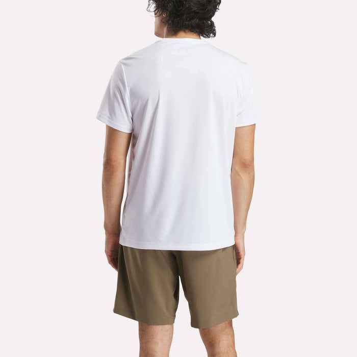 A person with short, dark curly hair stands with their back to the camera, wearing a white Reebok Men's ID Train Short Sleeve Tech Tee and olive green shorts against a light background.