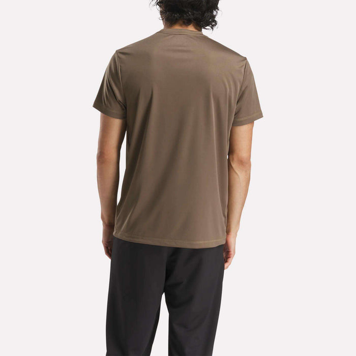 A person with dark wavy hair is seen from behind, wearing the Reebok Men's ID Train Short Sleeve Tech Tee in brown and black pants, set against a plain light background.