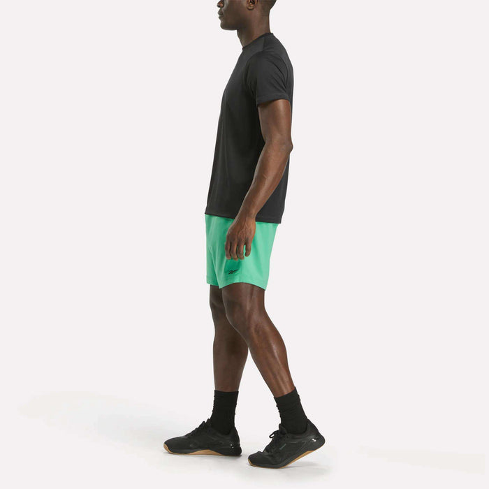 A man stands in profile wearing a black Reebok Men's ID Train Short Sleeve Tech Tee, green athletic shorts, black socks, and black sneakers against a plain white background.
