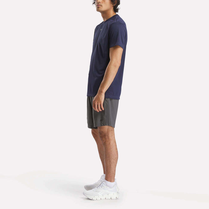 A man in profile wears a navy Reebok Men's ID Train Short Sleeve Tech Tee, gray shorts, and white athletic shoes against a plain white background.
