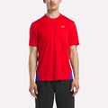 A man in a Reebok Men's Short Sleeve Vector T-Shirt in Vector Red/Breakout Blue and black shorts stands against a plain white background.