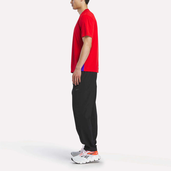 A man stands sideways against a plain white background, wearing a red Reebok Men's Short Sleeve Vector T-Shirt with RBK-DRY technology, black pants, and white athletic shoes with orange accents.
