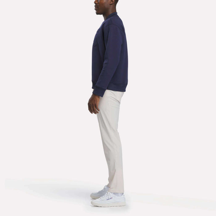 A person stands sideways against a plain white background, wearing the Reebok Men's Signature Golf Crewneck Sweatshirt in navy, paired with light-colored pants and white sneakers.