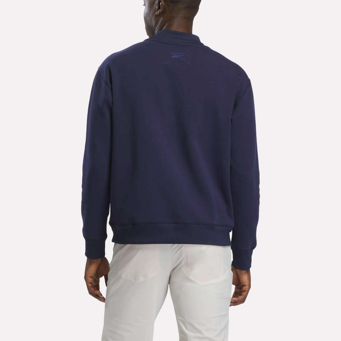 A man stands facing away, wearing a men's Reebok Signature Golf Crewneck Sweatshirt in navy blue featuring RBK-DRY technology, paired with light-colored pants against a plain white background.