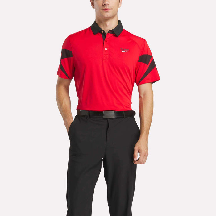 A man wearing a Reebok Men's Signature Vector Colorblocked Polo Shirt with black accents and black pants stands with one hand in his pocket against a plain white background.