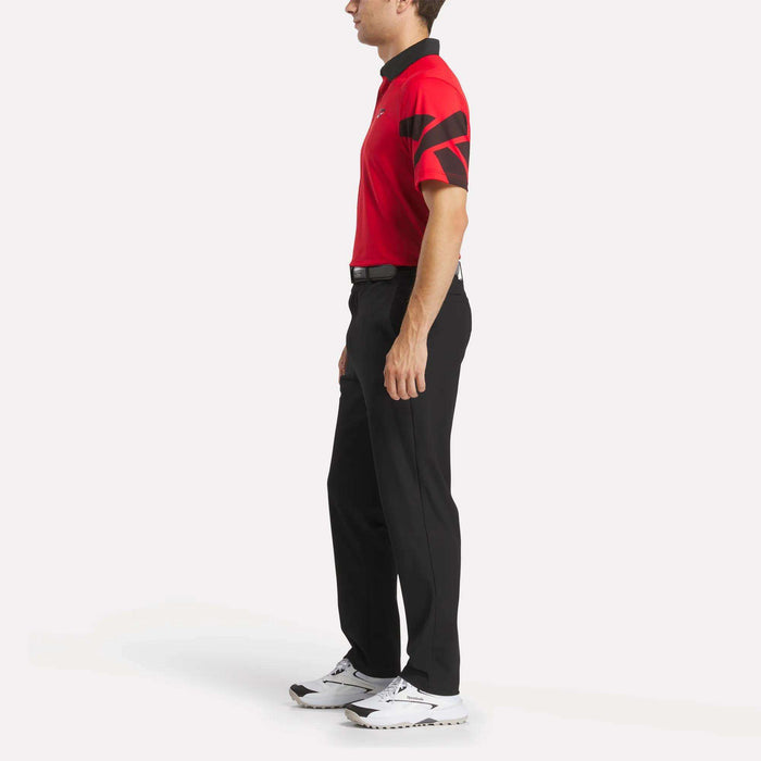 A man stands in profile facing left, wearing a Reebok Men's Signature Vector Colorblocked Polo Shirt with moisture-wicking, UPF 30+ protection, black pants, a black belt, and white athletic shoes against a plain white background.