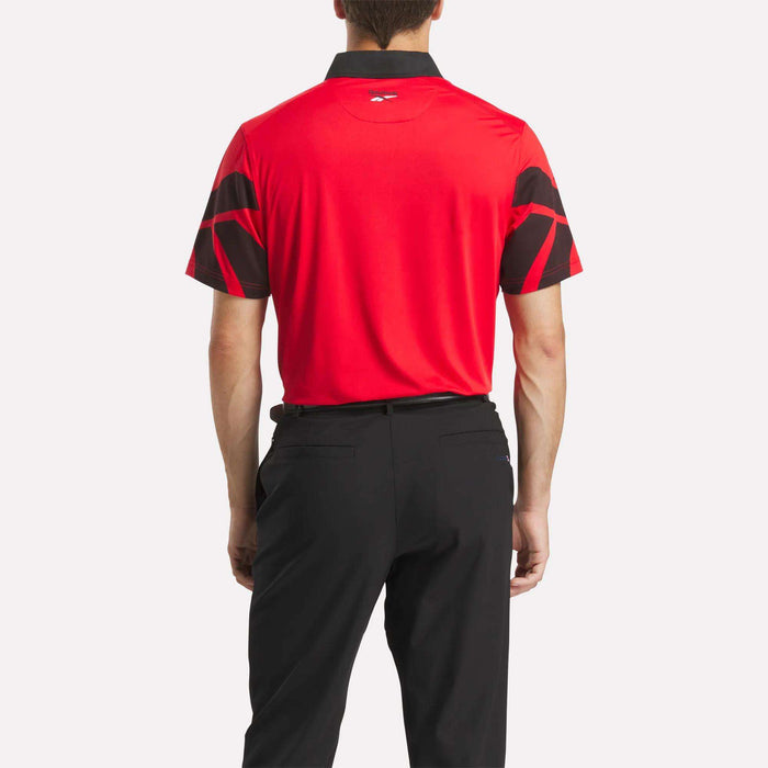 A man stands facing away, wearing the Reebok Men's Signature Vector Colorblocked Polo Shirt in red with black collar and patterned sleeves, featuring moisture-wicking fabric, paired with black pants against a plain light background.