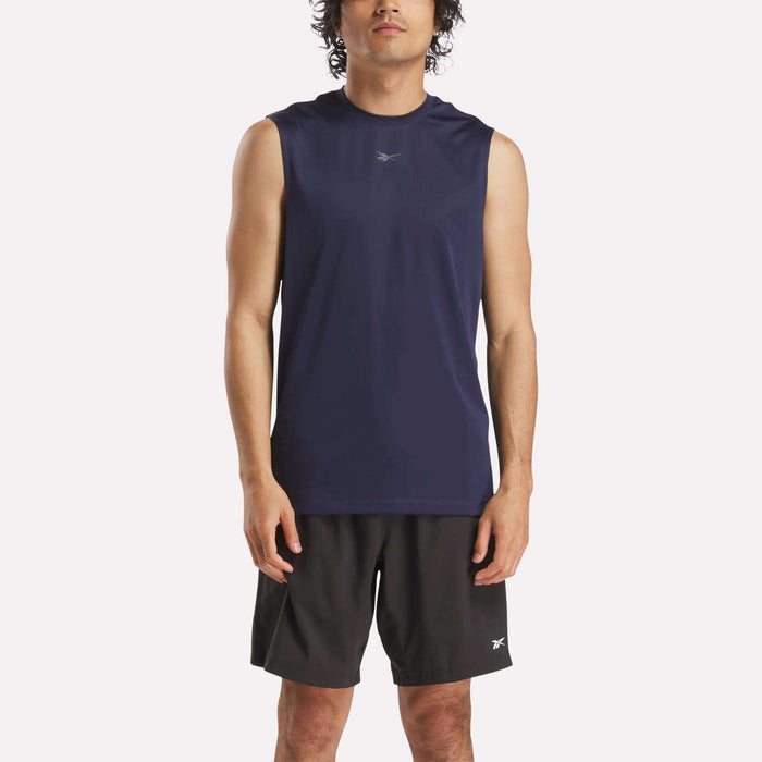 A person wearing a Reebok Men's Train Sleeveless Tech Tee in navy blue and black shorts stands facing forward against a plain light background.